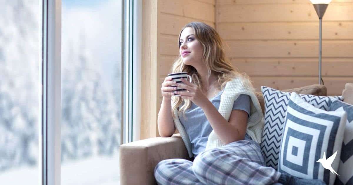 Blog-Previews A Woman Wearing Pajamas Sips A Hot Drink While Sitting On A Sofa, Looking Out At A Snowy Landscape Through A Large Window Featured On Our Windows And Doors Blog.