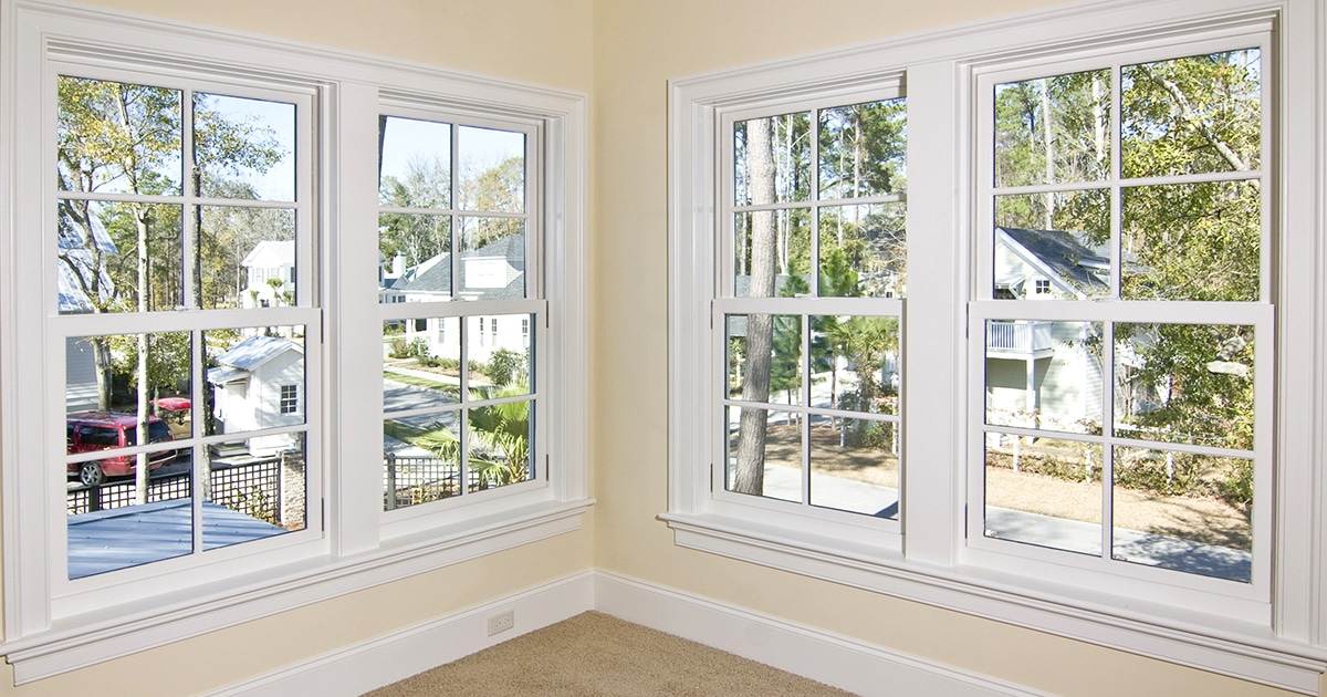 Doublehung-Windows Corner Of A Room With Two Sets Of White-Framed Double-Hung Windows, Perfect For Those Considering Buying Windows. These Overlook Neighboring Houses And Trees. Walls Are Light-Colored And The Floor Is Carpeted.