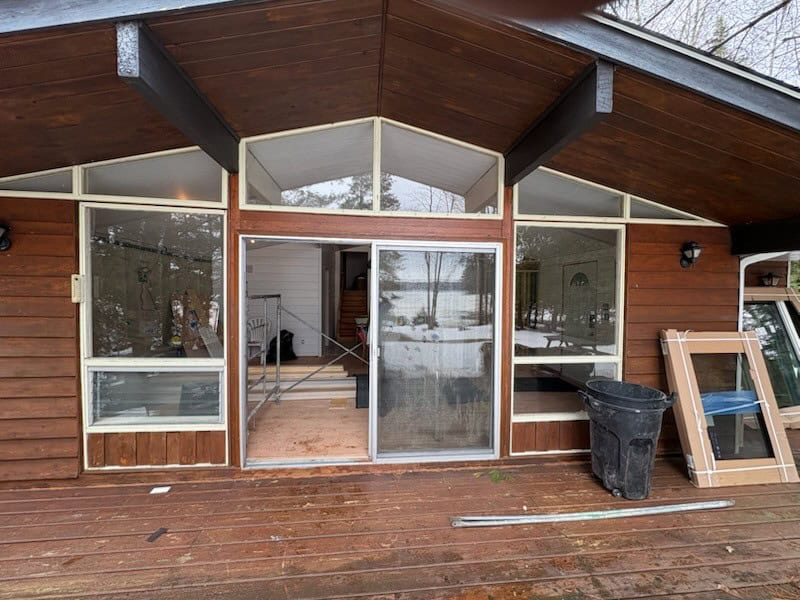 Before A Wooden Cabin With Large Windows And A Sliding Glass Door Leading To A Snow-Covered Interior. A Trash Bin And A Stacked Window Frame Are On The Wooden Deck.