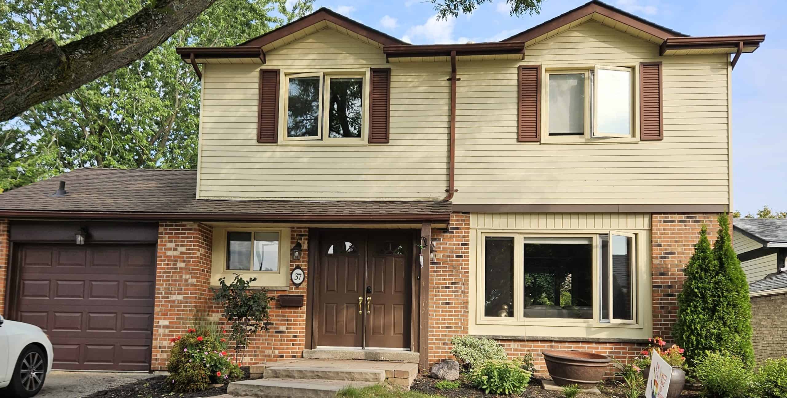Front Of House Two-Story Suburban House With Brick And Beige Siding, Brown Shutters, Double Front Doors, And Attached Garage; Landscaped Front Yard With Bushes And Flowers.