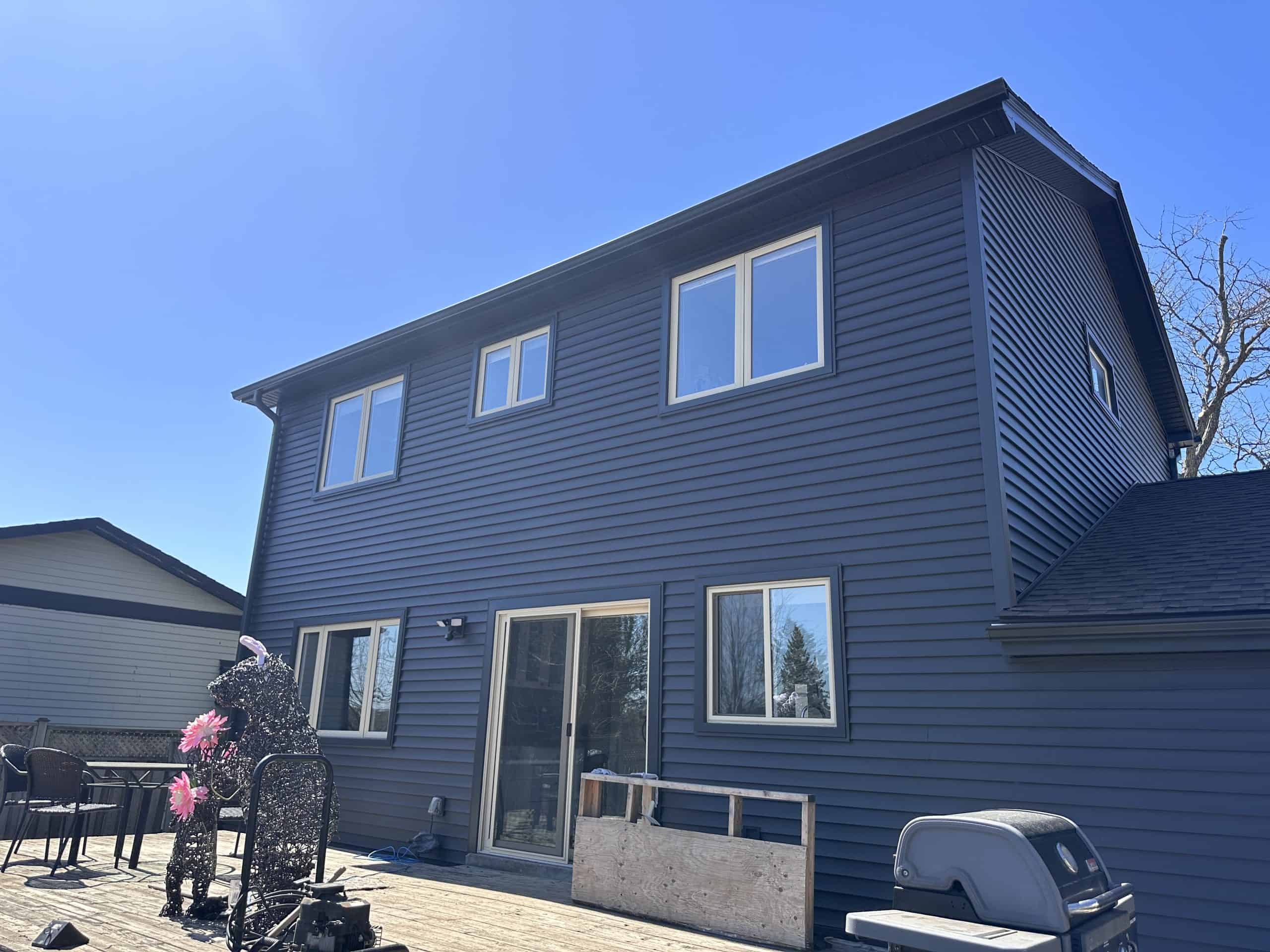 Img-4970 A Two-Story House With Dark Blue Siding, Several Windows, A Sliding Glass Door, A Grill, Patio Furniture, And A Metal Sculpture On A Wooden Deck Under A Clear Blue Sky.