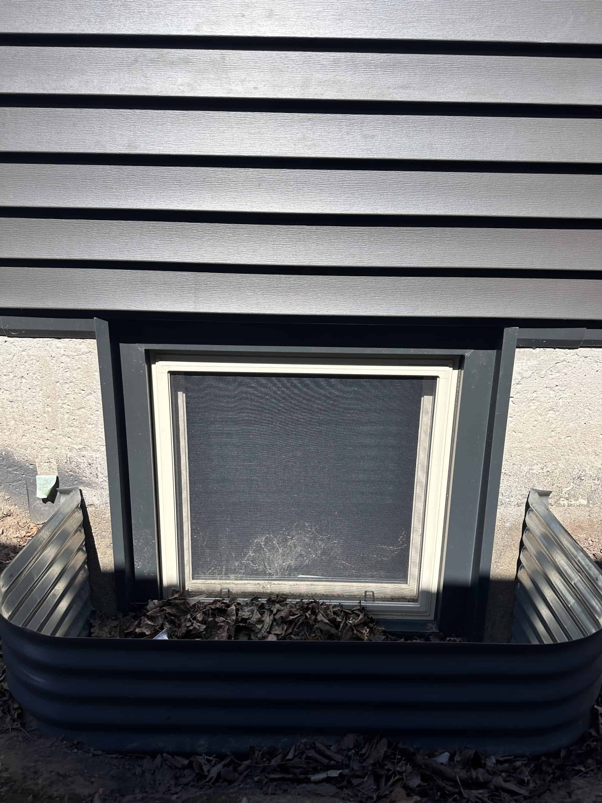 Img-4977 A Basement Window With A Metal Window Well, Surrounded By Dark Horizontal Siding And A Concrete Foundation, With Leaves Collected At The Bottom.
