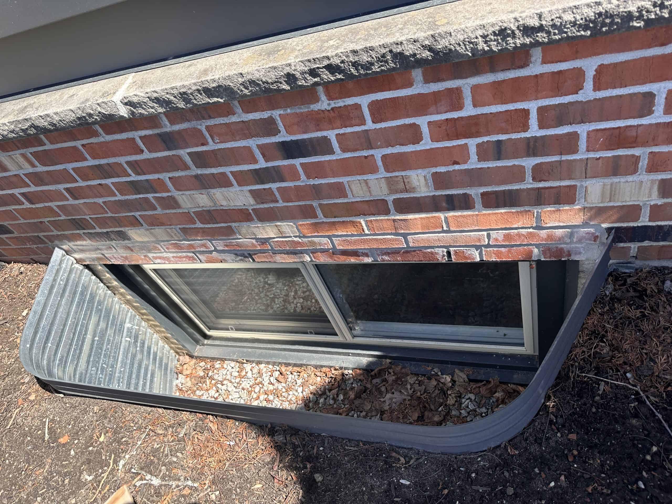 Img-4978 A Ground-Level Basement Window With A Metal Window Well, Scattered Leaves And Debris Inside, Set In A Brick Wall With A Concrete Ledge Above.