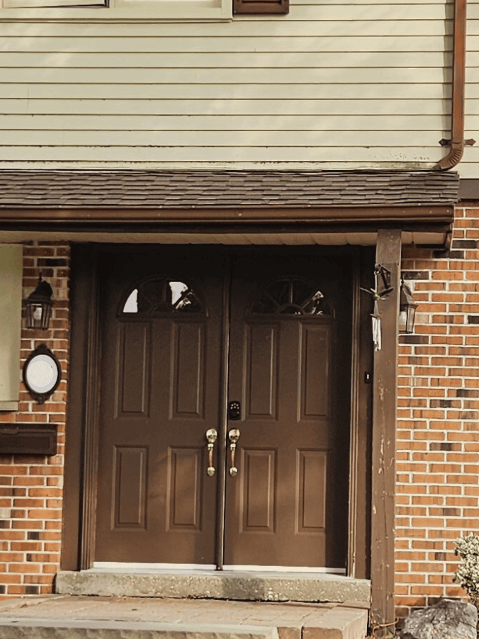 Untitled Design (17) A Pair Of Brown Double Front Doors With Gold Handles, Set Beneath A Small Awning, Next To A Brick Wall And Lantern-Style Lights.