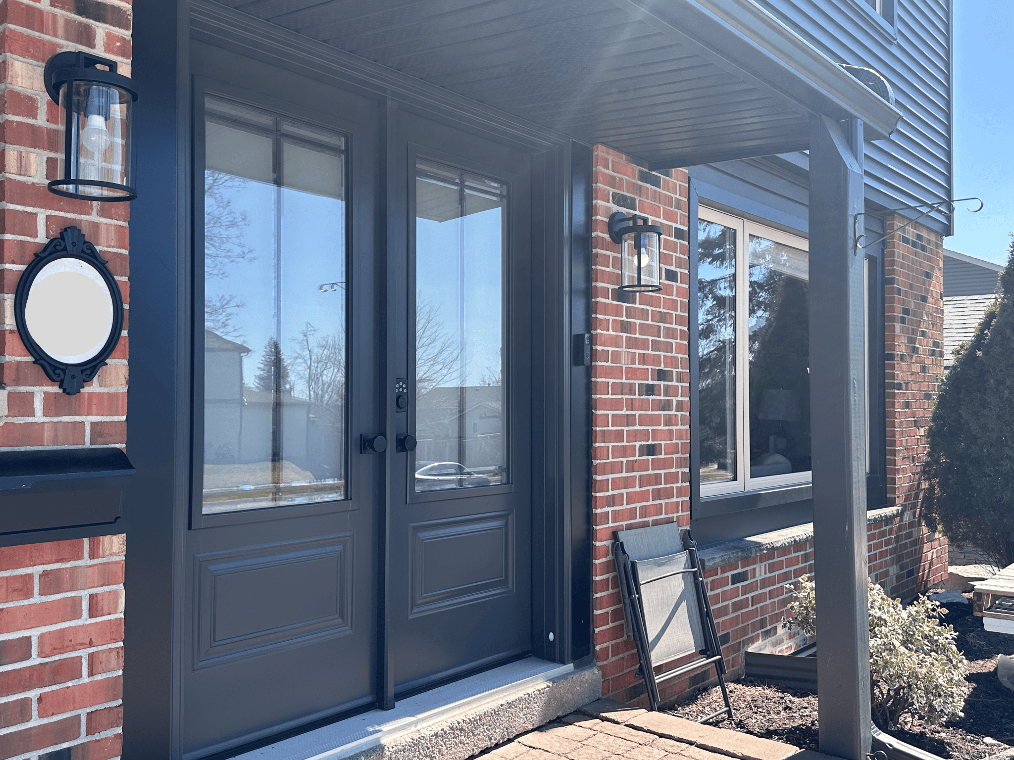 Untitled Design (18) A Double Glass Front Door With Dark Trim On A Red Brick House, Flanked By Wall-Mounted Lantern Lights And A Small Garden Bed With Shrubs.