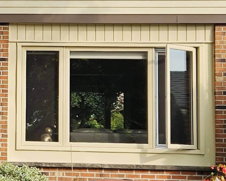 C39641C8-845D-4F27-9D7F-Efca283E9A2F A Beige-Framed Window Set In A Brick Wall, With One Side Window Panel Open. Trees And Part Of A House Roof Are Visible Through The Glass.