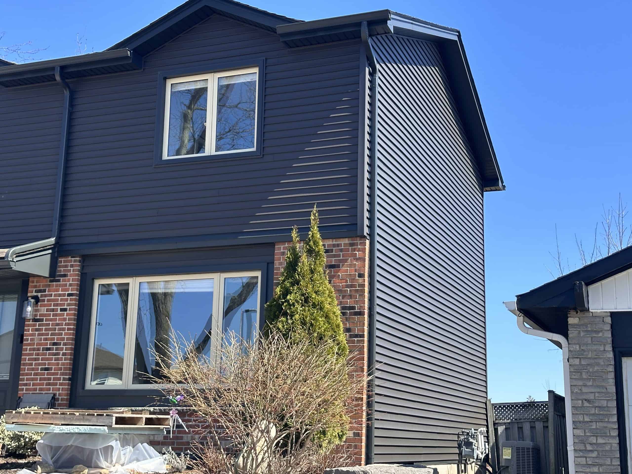 Crop On The Side A Two-Story House With Dark Siding And Red Brick On The Lower Level, Featuring Large Windows And A Small Shrub In The Front Yard Under A Clear Blue Sky.