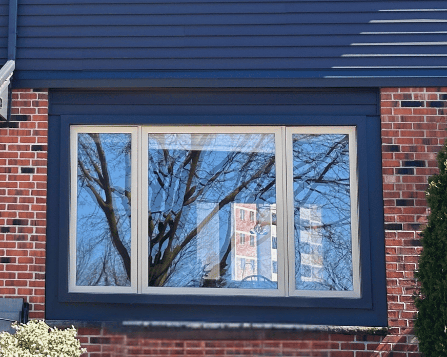 F9Eb7600-E428-4Da9-9780-01990086B218 A Large Window On A Brick House Reflects Leafless Tree Branches And Sky; Part Of A Building And Objects Are Visible Inside Through The Glass.